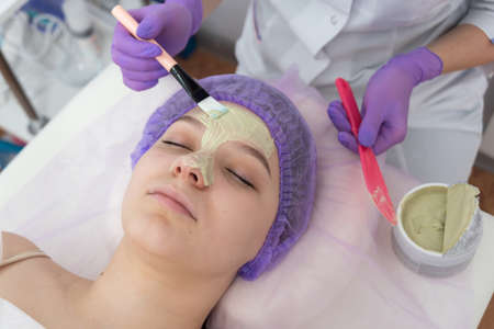 In the beauty salon, the beautician puts a mask of green clay on the face of a young girl. Daily...