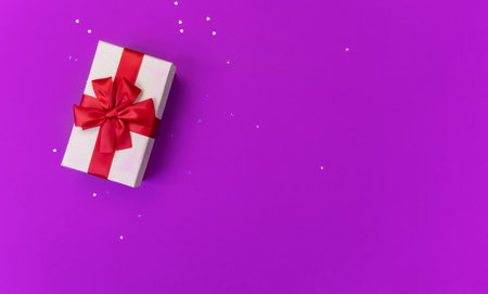 Gift box with red bow on purple background with hearts confetti. Top view.