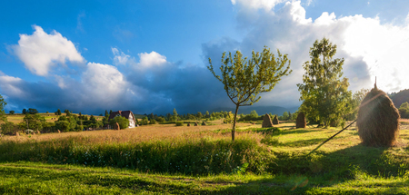 Rural landscape with haystacks in a summer sunny day. Rural mountain landscape with storm clouds. Thunderstom under field. House in the field of golden wheat.の写真素材