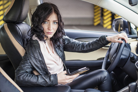 Beautiful sexy female model in leather clothes with a white car and a smartphone in the Parking lotの写真素材