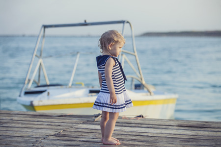 Little baby girl in sailor dress on wooden pier on the background of the boatの写真素材