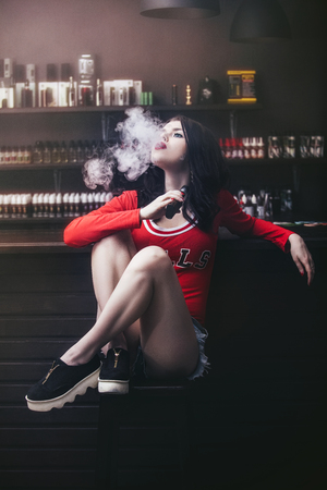 Young beautiful brunette woman with fashion makeup at the bar with a with vapor from electronic cigaretteの写真素材