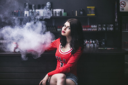 Young beautiful brunette woman with fashion makeup at the bar with a with vapor from electronic...