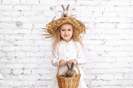 Beautiful child girl with curly hair and fluffy rabbits animals sitting on a hatの写真素材