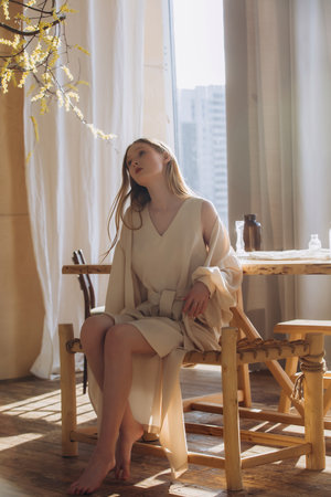 Beautiful blonde model, young girl, teenager in the studio in a beige dress in the interior with a tableの写真素材