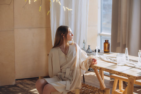 Beautiful blonde model, young girl, teenager in the studio in a beige dress in the interior with a tableの写真素材
