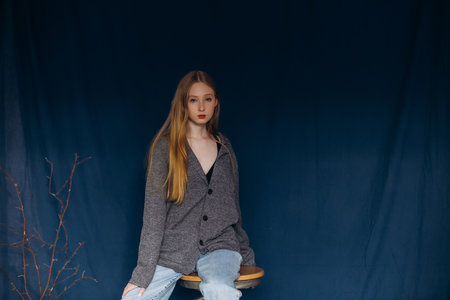 Blonde model, young girl, teenager in the studio in a jacket and jeans on a chair on a blue...