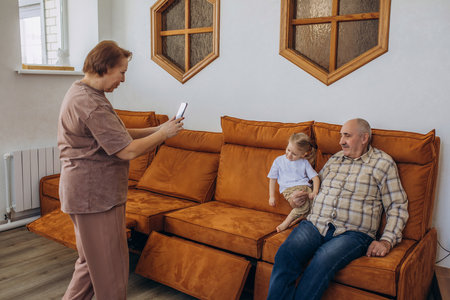 Grandmother takes a photo of her grandfather and baby granddaughter sitting on the couch at home on her smartphoneの写真素材