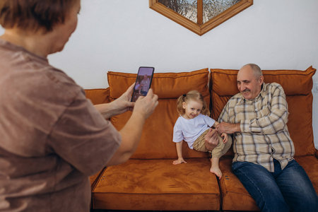 Grandmother takes a photo of her grandfather and baby granddaughter sitting on the couch at home on her smartphoneの写真素材