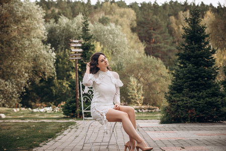 Beautiful young brunette woman in white dress on outdoors in the park sitting on a chairの写真素材