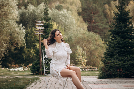 Beautiful young brunette woman in white dress on outdoors in the park sitting on a chairの写真素材