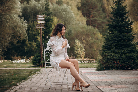 Beautiful young brunette woman in white dress on outdoors in the park sitting on a chairの写真素材