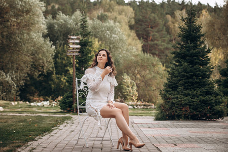 Beautiful young brunette woman in white dress on outdoors in the park sitting on a chairの写真素材