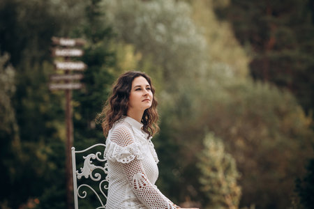 Beautiful young brunette woman in white dress on outdoors in the park sitting on a chairの写真素材