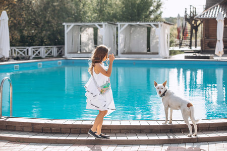 Beautiful baby girl in a white dress with a dog, by the pool outsideの写真素材