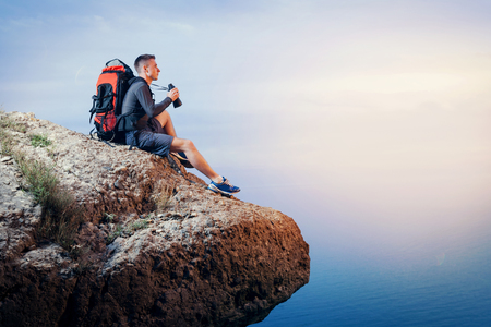 A young man hiker  with a backpack hiking looking through binoculars sitting on a rock mountain over the sea.の写真素材