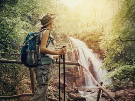 Hiking. Hikers woman with a backpack and a hat looking at a waterfall in the forest.の写真素材