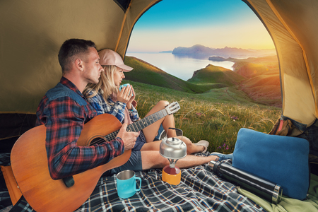 Happy backpackers with a guitar from the tent look at the mountains. Campsite. People in camping. Young couple in a tent enjoys the sunset and nature. View from inside.の写真素材