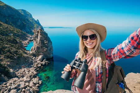 Young woman traveler in sunglasses with binoculars makes selfie sea view. Portrait of a happy girl...
