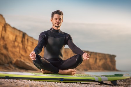 Young man meditating in lotus position on a SUP board on sea beach. Yoga, on a paddle board outdoor.の写真素材