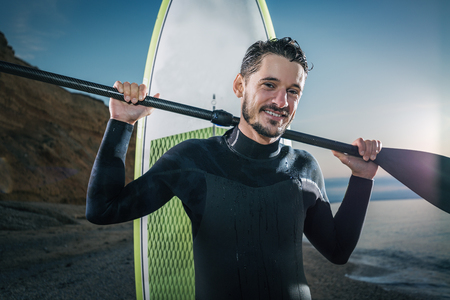 Portrait of a surfer with SUP board on the beach. Young man with a stand up paddleboard at sunset. Extreme sport concept. Male surfer lifestyle.の写真素材