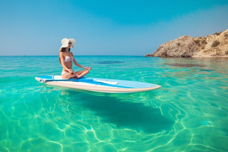 Young beautiful woman meditating in a sea at SUP paddleboarding. Healthy lifestyle. Girl in yoga...