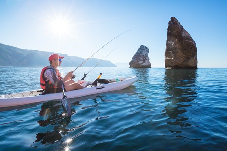 A man fishing on a kayak boat in the sea near the rocks at the shore of island mountain. Young fisherman kayaking.の写真素材