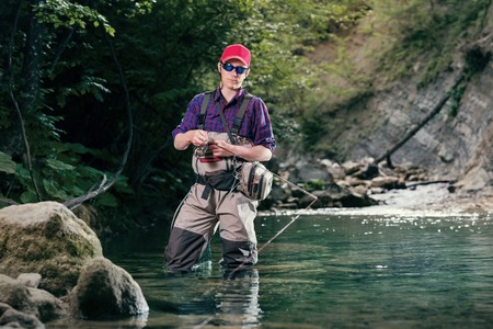 A fisherman chooses bait for trout fishing in the river. Fishing in the creek fly fishing lure. Activity sports fishing wading.の写真素材