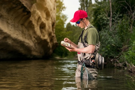A fisherman chooses bait for trout fishing in the river. Fishing in the creek fly fishing lure. Activity sports fishing wading.の写真素材