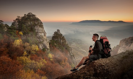 Young man hiker with backpack and binoculars sitting on a cliff and enjoying a Mountain view.の写真素材