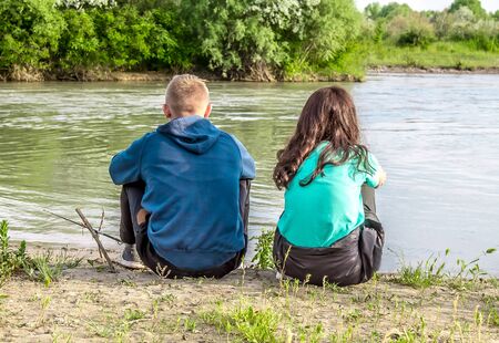 Young boy and girl are sitting on the ground by the riverの写真素材