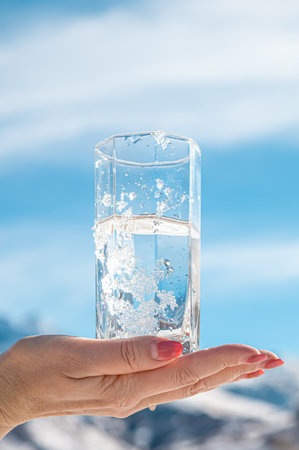 Glass cup with cold water in hand on a background of sky and mountains in the snow.の写真素材