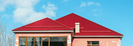 The roof of a cottage with a metal coating, snow guards, a chimney, against the background of a blue sky. Inclined roof of a residential building made of metal profiled sheet, metal slate.の写真素材