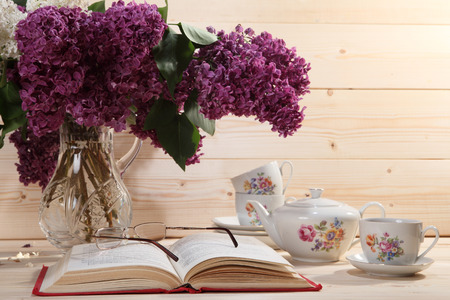 Bouquet of lilacs, open book, spectacles, teapot and cup of tea on wooden backgroundの写真素材