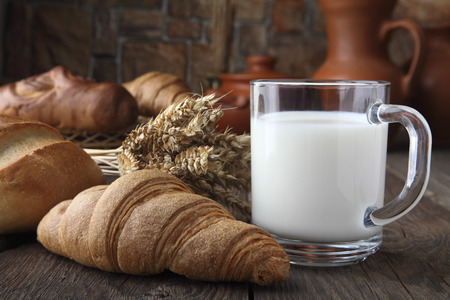 Glass mug with milk, bun and bakery products lying on an old table on the background of wheat spikes and ceramic dishes. Close-upの写真素材