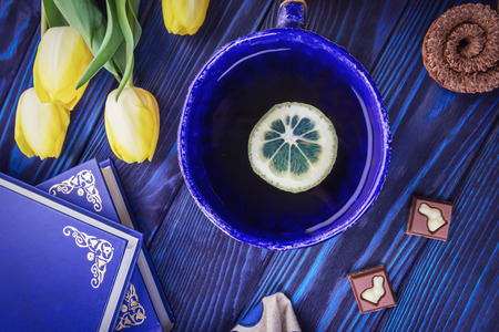 Tea with lemon, books and spring yellow tulips on a blue wooden background. Chocolate with a bunny figurineの写真素材
