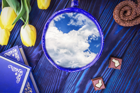 Cup with beautiful sky inside, books and spring yellow tulips on a blue background. Concept better weather forecast for spring and Easterの写真素材