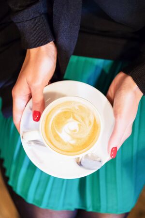 Beautiful female hands holding a white cup of coffee cappuccino. Manicure with red nail polish. Top view, verticalの写真素材
