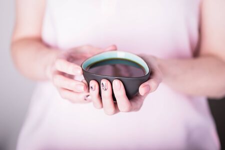 Beautiful female hands holding black turquoise cup of coffee on pink dress background. Manicure with pink color nail polish, floral monstera leaves patternの写真素材