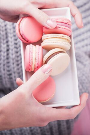 Female hands holding rectangular plate with delicious pink and beige macaroons or macarons on pink and gray background. Top view, copy spaceの写真素材