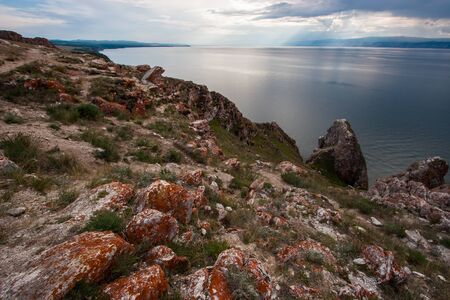 Stones with red moss on the rocks of Lake Baikal on Olkhon Island. Between the stones green grass. Mountains behind the lake. Clouds in the sky. Sun rays shine through the clouds.の写真素材