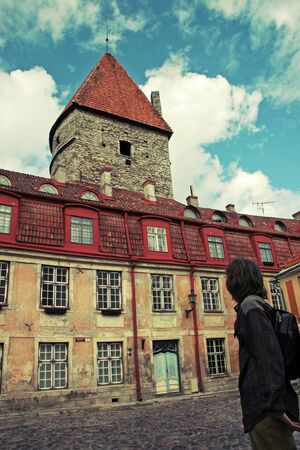 A man stands with his back and looks at an old beautiful building with red tiles in the old city of Tallinn. The walls are yellow shabby. Blue door. Beautiful old tower of limestone. The sky is blue with clouds. Tinted in yellow.の写真素材