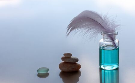 A bottle of blue scented oil and a sticking bird feather. A pyramid of stones and a piece of glass is folded. Blue blurred background. Place for text.の写真素材