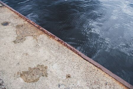 The edge of the old concrete pier with a rusty iron edging. Diagonal in the frame. Blue water with ripples and a reflection of the sky.の写真素材