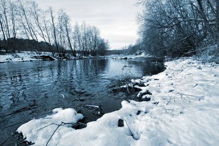River without ice in winter with snow on the shore and growing trees. Water with ripples and reflections of trees. Lots of snow on the shore. Toning in blue. There is grass in the water. The sky is gray with clouds without the sun.の写真素材