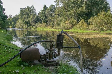 A big old water pump stands on the bank of the river and pumps water. The river is overgrown with grass. Trees grow on the shore.の写真素材