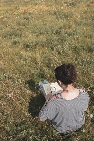 A girl tourist in a striped sweater sits in a field among the grass and looks at a map through a...