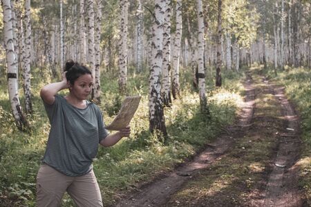A pensive girl looks at a map in the forest. Verifies the route. Birch forest with a forest road. Girl in a striped sweater and light pants. Sunny. Selective focus on the map. The background is blurry.の写真素材