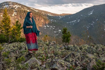 Beautiful cute girl in a dress and a scarf stands on large stones in the mountains. Against the background of mountains with snow overgrown with forest. Selective focus on the girl.の写真素材