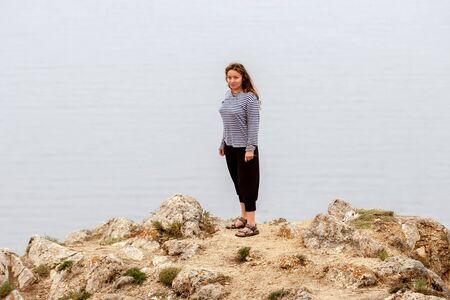 A surprised beautiful girl stands on a rock above the water. European with long dark hair. Singlet, bodysuit and sandals.の写真素材